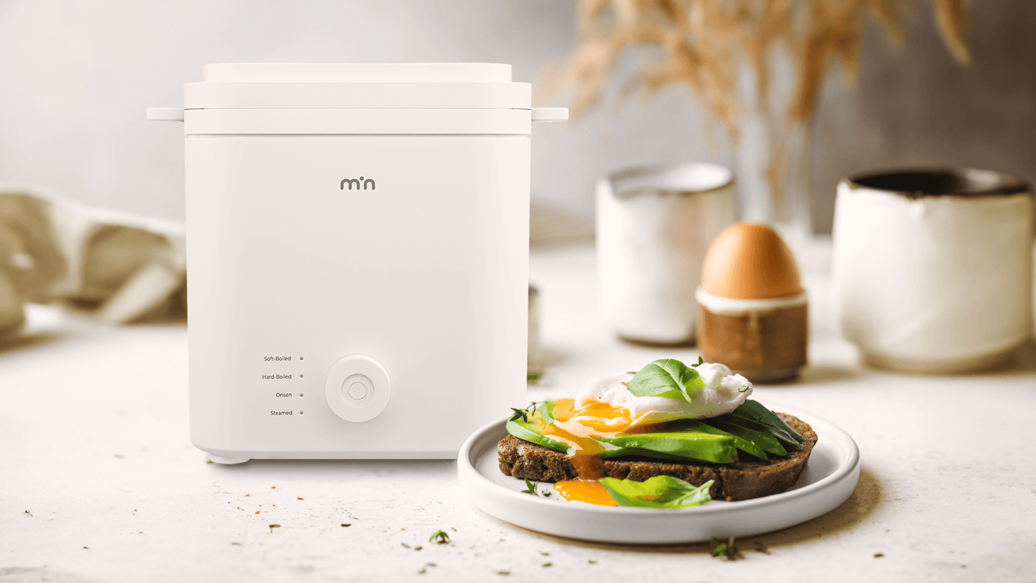 min Egg cooker placed on a minimalist background, next to a breakfast plate featuring toast and perfectly cooked eggs.